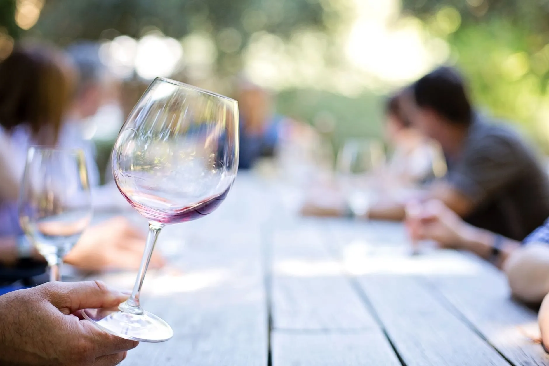 A hand holding a wine glass at a long outdoor tasting table, guests blurred behind
